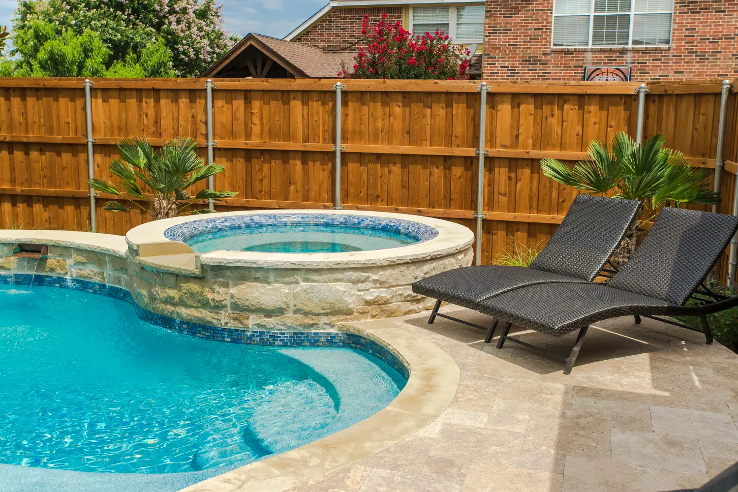 Freeform pool with a raised spa and travertine deck in a fenced backyard, built by Hobert Pools & Spas