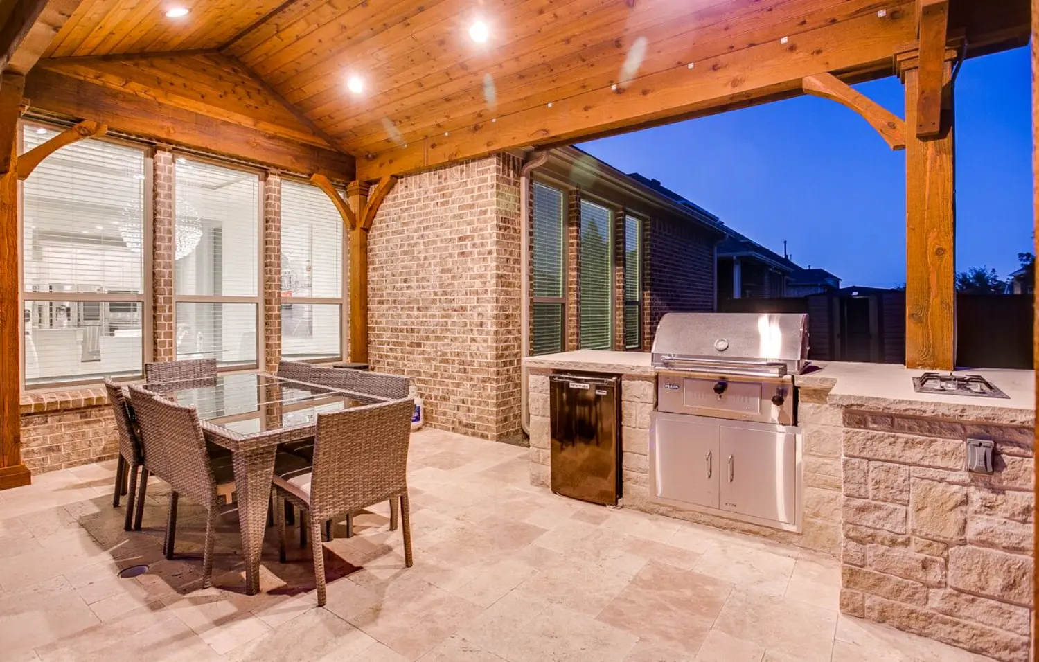 Covered outdoor kitchen with a built-in grill and dining area on a travertine patio, built by Hobert Pools & Spas